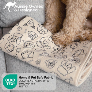 A beige pet blanket with black line drawings of dog faces, potty training symbols, and paw prints, shown on a gray sofa with a fluffy light brown dog's legs visible. Text overlays include 'Aussie Owned & Designed', 'Home & Pet Safe Fabric', and OEKO-TEX certification details.
